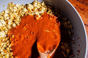 Crumbled tofu in a pan with savory sofritas red sauce is being stirred with a wooden spoon on a wooden surface.