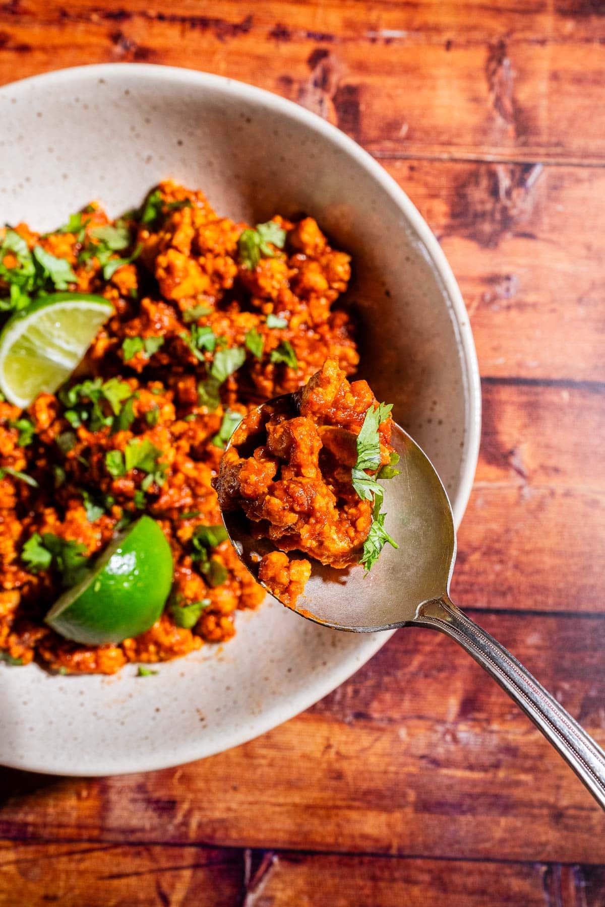 A spoonful of spicy sofritas minced dish garnished with chopped cilantro and lime wedges in a bowl on a wooden table.