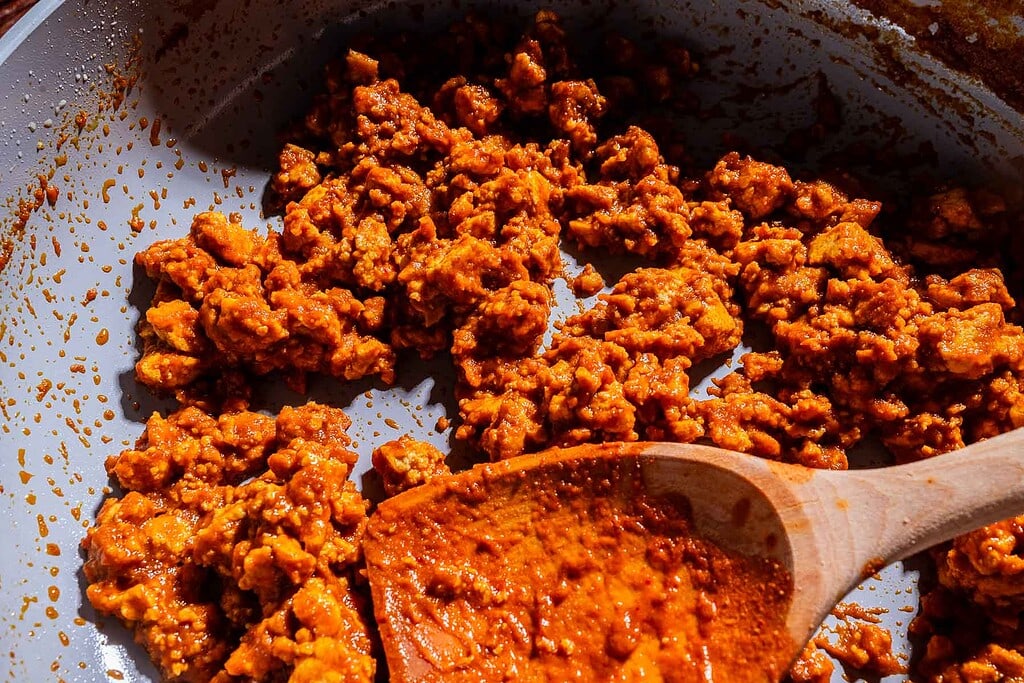 Cooked ground meat in red sauce, reminiscent of sofritas, being stirred with a wooden spoon in a skillet.