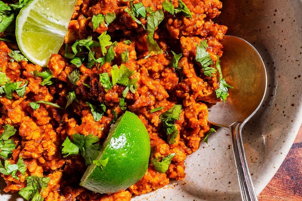 A bowl of seasoned sofritas topped with chopped cilantro, served with lime wedges and a spoon on the side.
