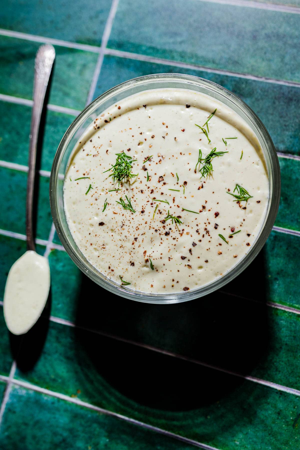 A glass bowl of creamy tofu ranch sauce garnished with black pepper and fresh dill sits on a green tiled surface beside a spoon.