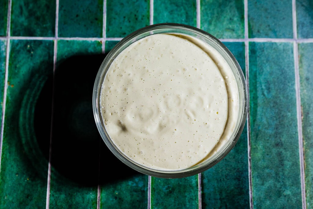 A glass jar filled with bubbly, creamy sourdough starter sits on a green tiled surface, beside a small bowl of tofu ranch.