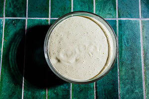 A glass jar filled with bubbly, creamy sourdough starter sits on a green tiled surface, beside a small bowl of tofu ranch.