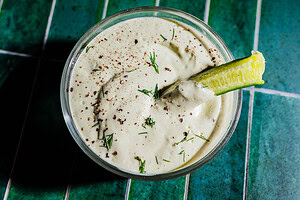 A glass bowl of creamy tofu ranch dip with fresh dill and black pepper, served with a pickle spear, on a green tiled surface.