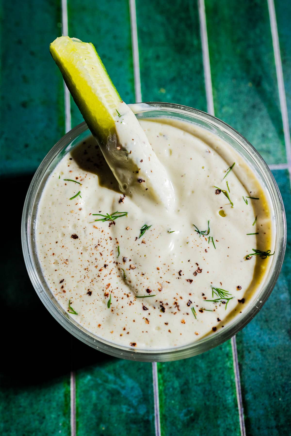 A glass of creamy tofu ranch dip garnished with herbs and black pepper, with a pickle spear partially submerged, on a green tiled surface.
