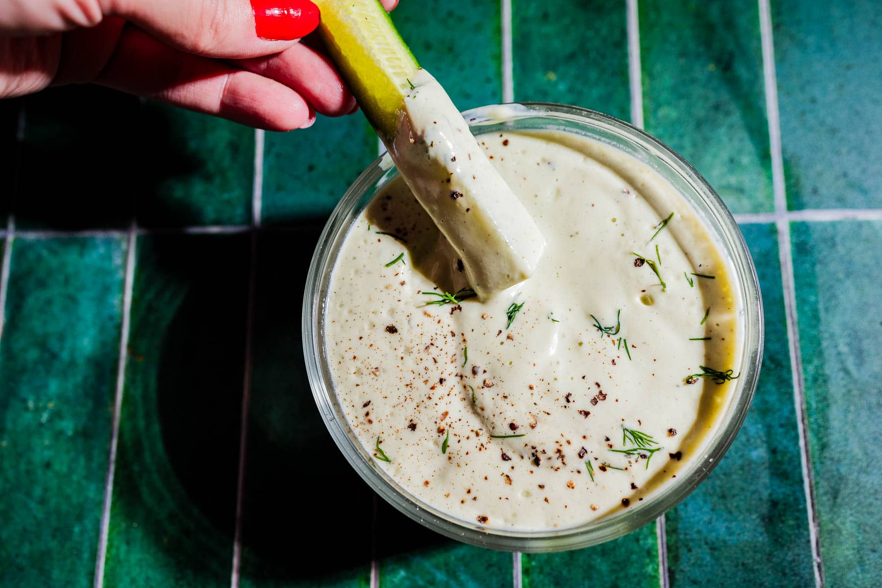 A hand dips a pickle spear into a bowl of creamy tofu ranch sauce, garnished with herbs and cracked black pepper, on a green tiled surface.