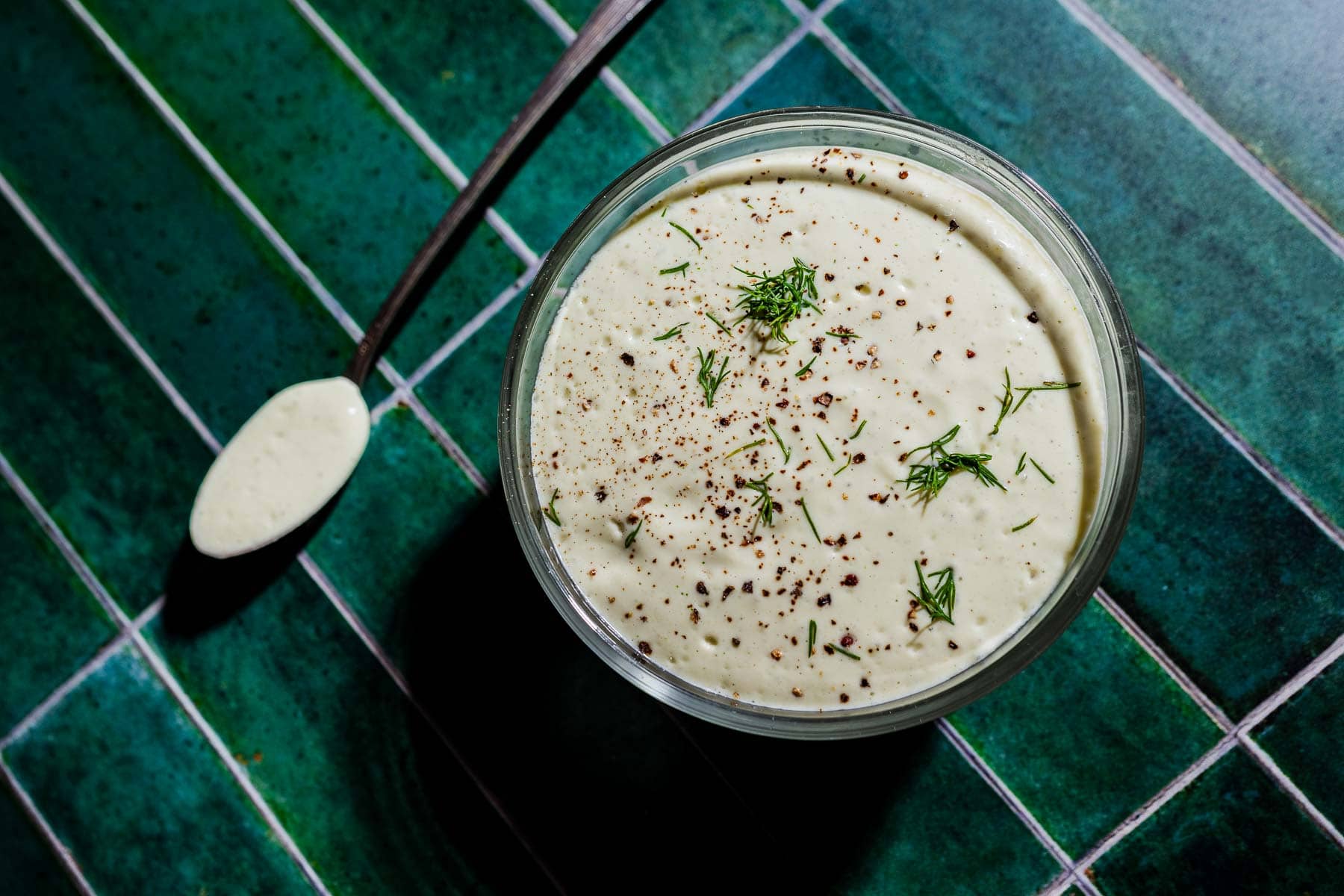 A glass bowl of creamy tofu ranch dip garnished with dill and pepper sits on a green tiled surface, with a spoonful of dip beside it.
