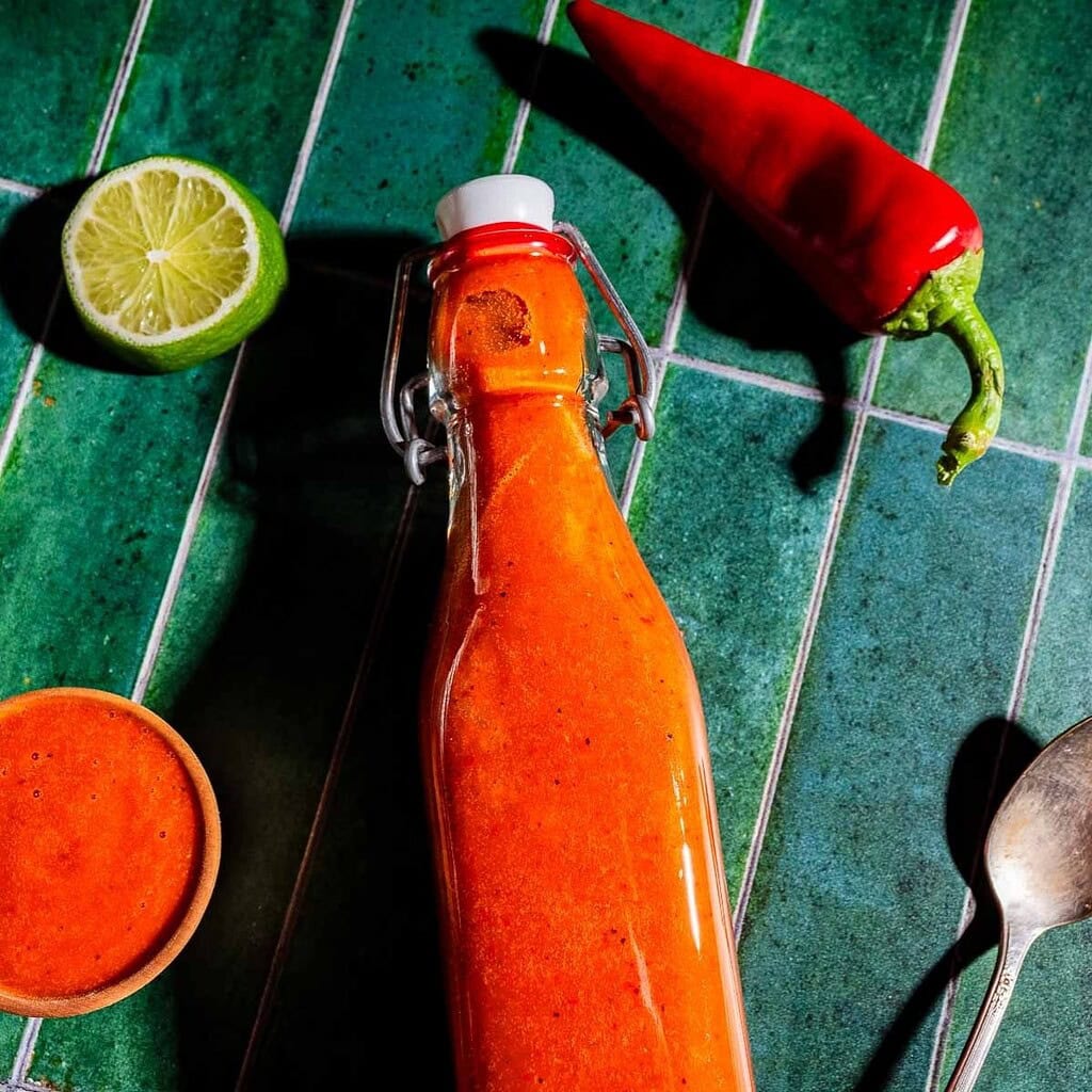A bottle of red hot sauce is placed on a green tiled surface, with a red chili pepper, a halved lime, a small bowl of sauce, and a spoon nearby.