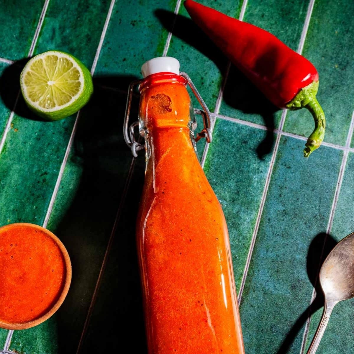 A bottle of red hot sauce is placed on a green tiled surface, with a red chili pepper, a halved lime, a small bowl of sauce, and a spoon nearby.