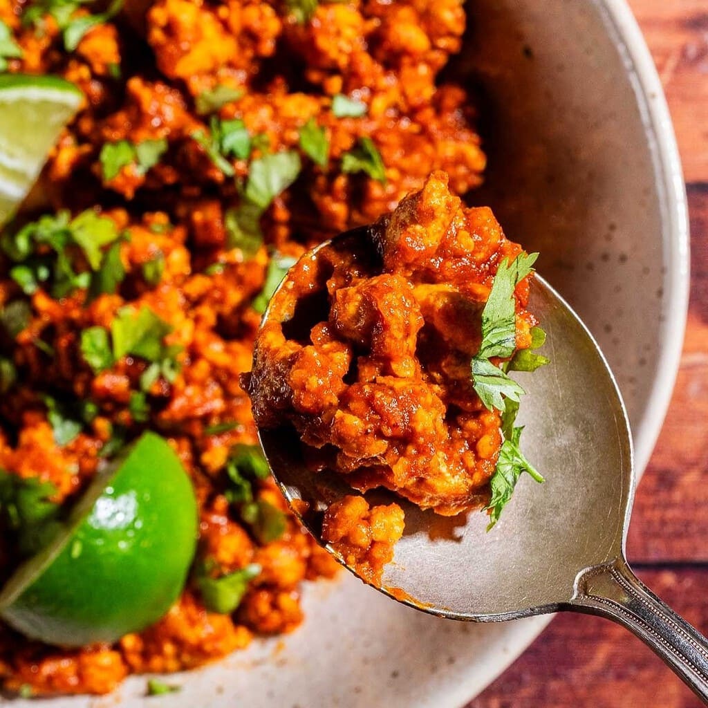 A close-up of a spoonful of seasoned sofritas ground meat garnished with chopped cilantro, with lime wedges visible in the bowl.