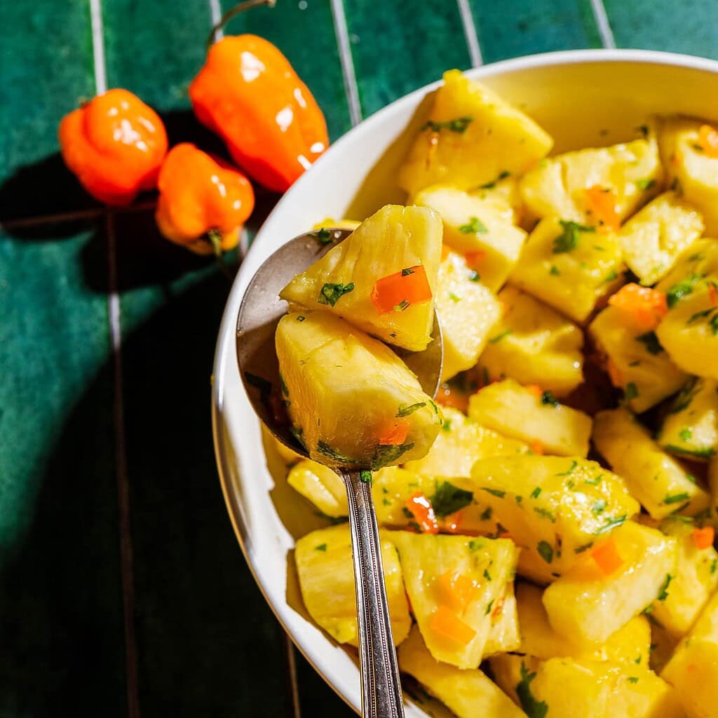 A bowl of pineapple salad with herbs and chopped peppers, with a spoon and three whole orange peppers on a green striped surface.