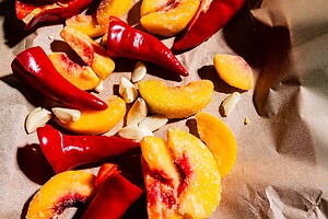 Red chili peppers, peach slices, and garlic cloves on brown parchment paper in bright lighting.