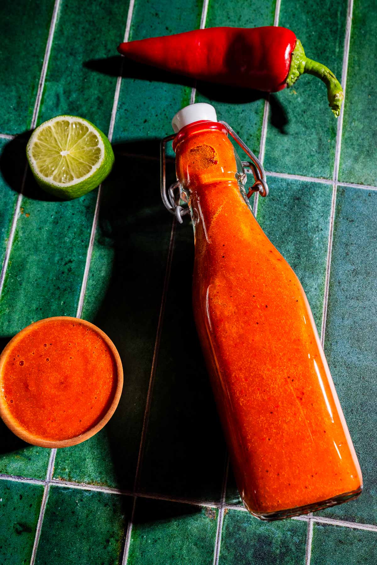 A glass bottle of red hot sauce sits on green tile with a red chili pepper, half a lime, and a small bowl of sauce nearby.