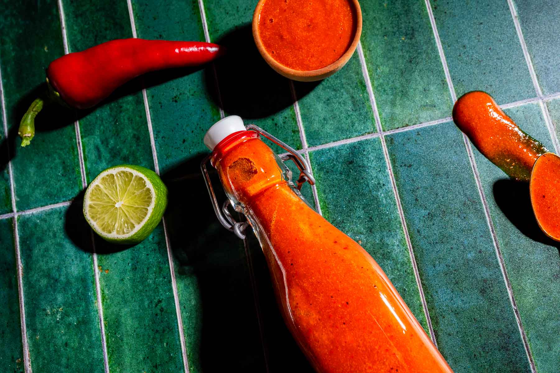 A bottle of red hot sauce lies on green tiles, surrounded by a halved lime, a red chili pepper, a small bowl of sauce, and a spoon with spilled sauce.