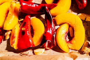 Sliced peaches, red chili peppers, and garlic cloves on parchment paper, brightly lit, arranged for roasting or cooking.
