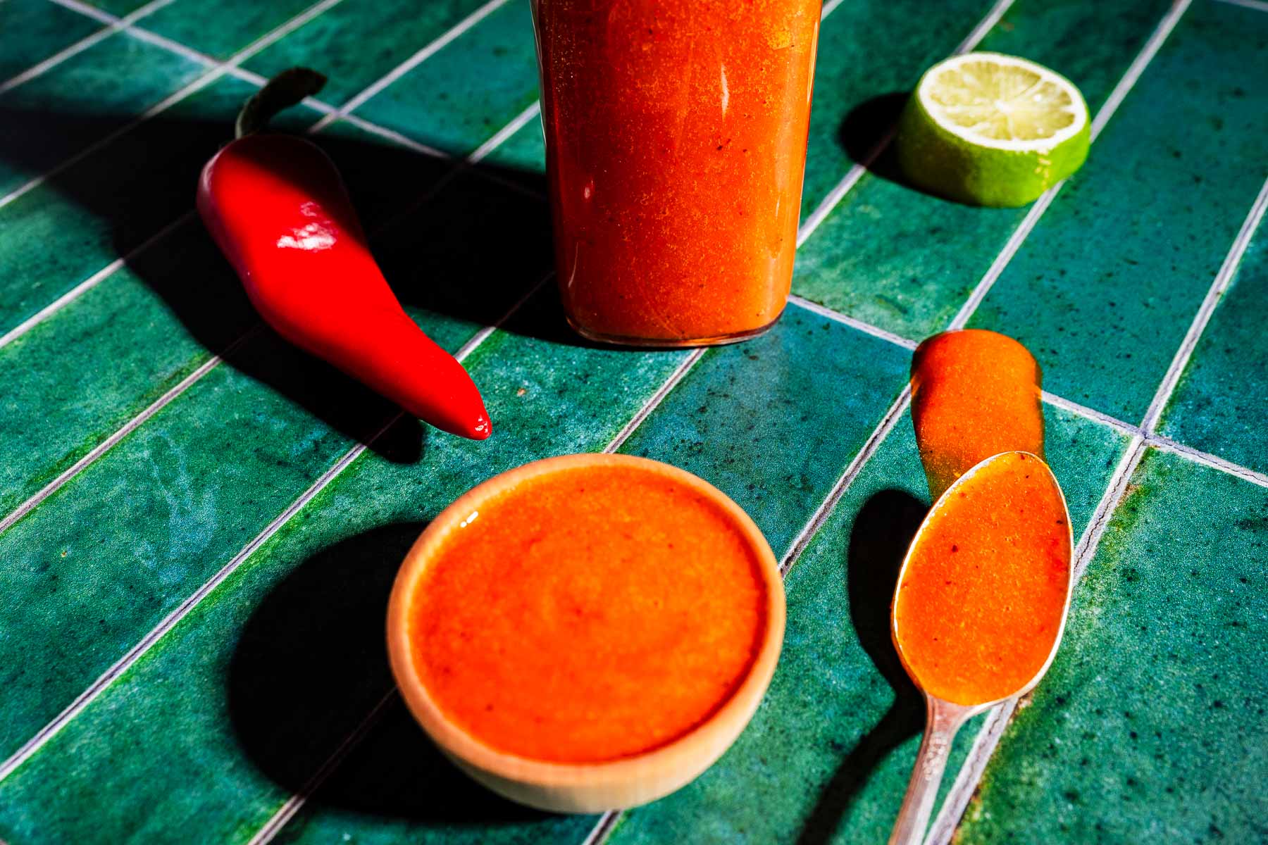 A bowl, bottle, and spoon of red chili sauce sit on green tiles with a red chili pepper and a halved lime nearby.