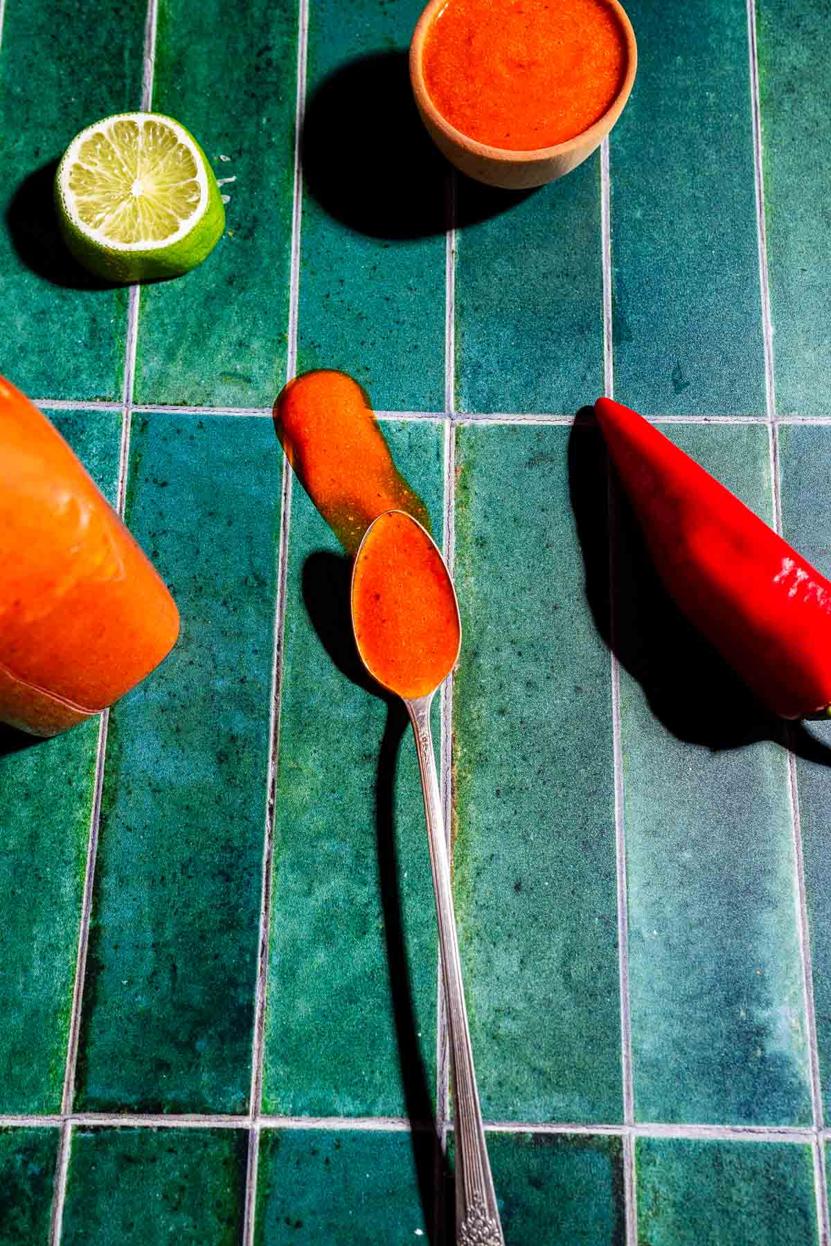 A spoon with red sauce, a spilled streak of sauce, a red chili pepper, a halved lime, and a small bowl of sauce on a green tiled surface.