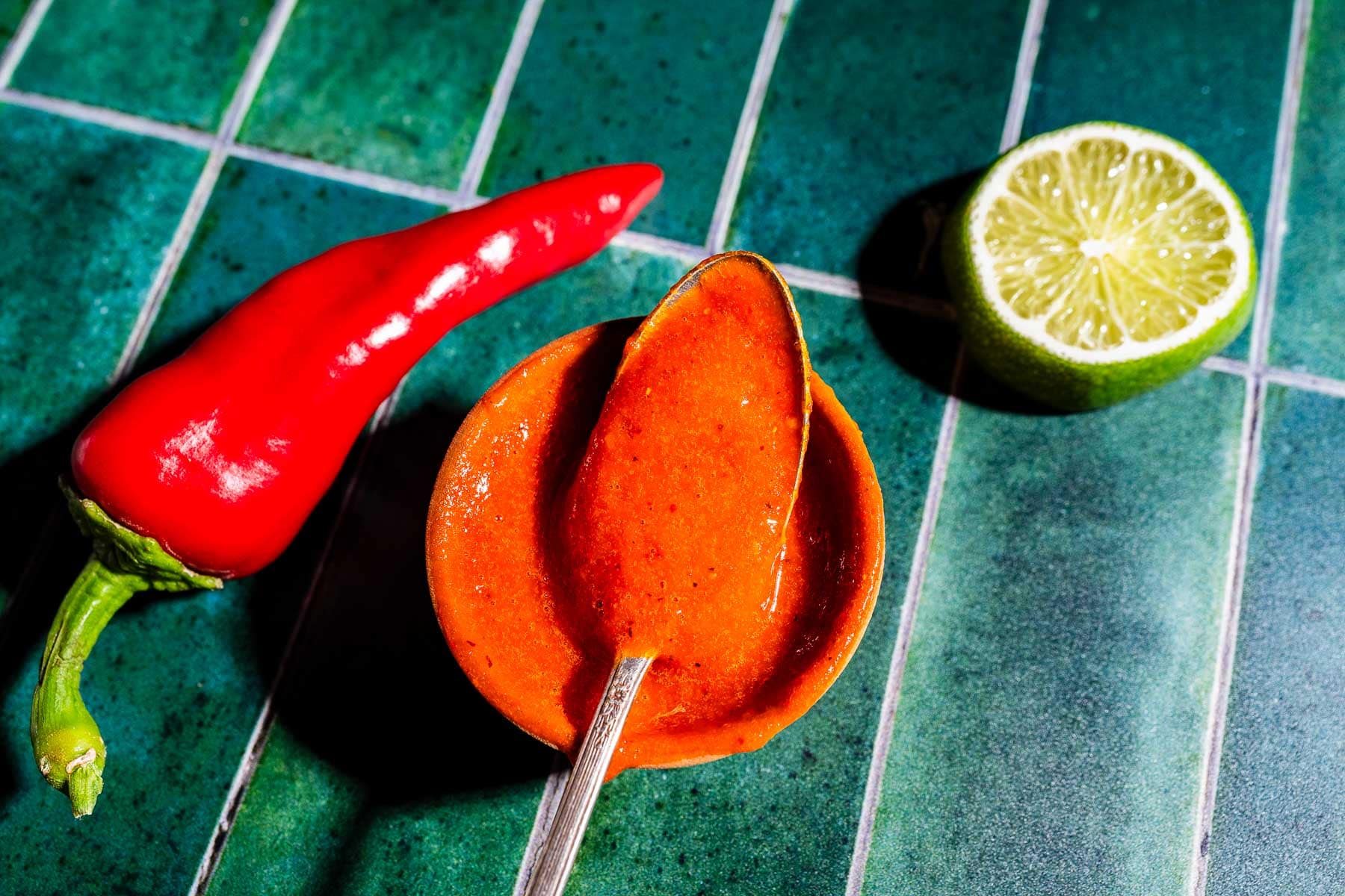 A red chili pepper, a halved lime, and a spoonful of red sauce in a small bowl are arranged on a green tiled surface.