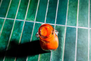 A glass bottle filled with orange-red sauce sits on a green tiled surface, viewed from above.
