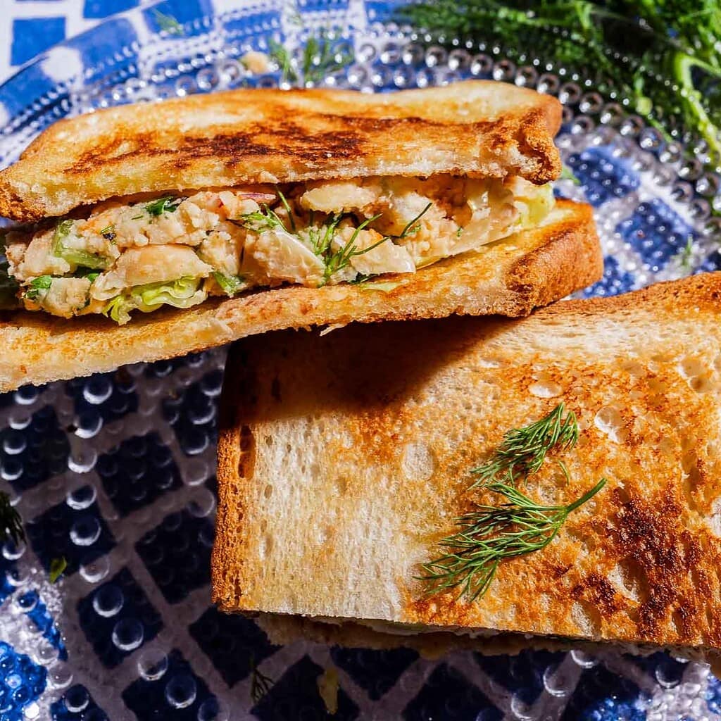 A toasted butter bean salad sandwich, filled with chicken salad and lettuce, is cut in half and served on a blue and white patterned plate, garnished with fresh dill.