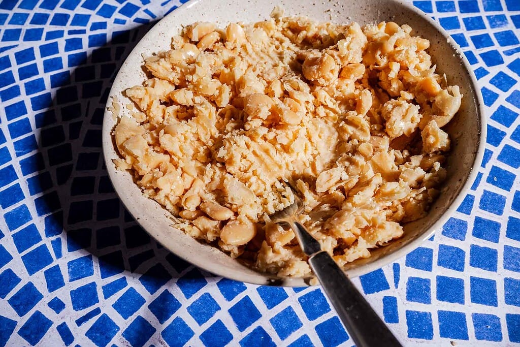 A bowl filled with mashed white beans for a butter bean salad sandwich sits beside a fork on a blue and white tiled surface.