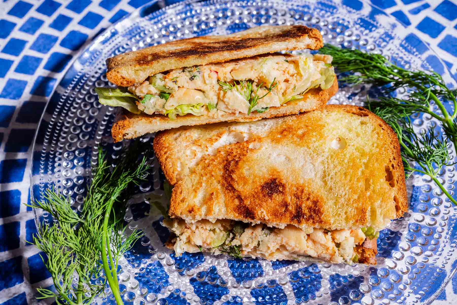 A grilled butter bean salad sandwich filled with a creamy mixture, cut in half and served on a clear plate, garnished with fresh dill on a blue mosaic surface.