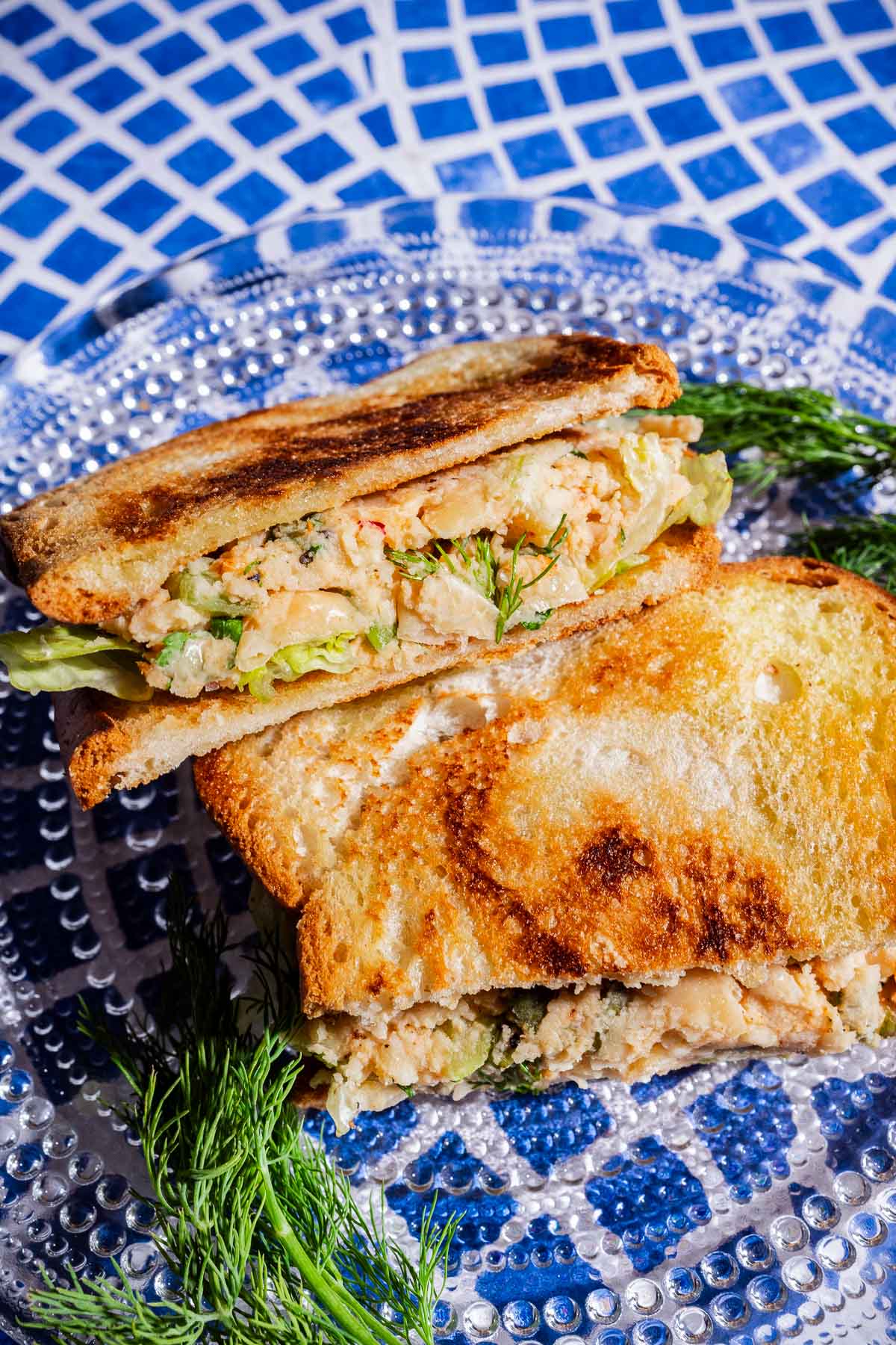 A toasted butter bean salad sandwich or classic chicken or tuna salad mixture, garnished with fresh dill, served on a clear glass plate with a blue and white mosaic background.