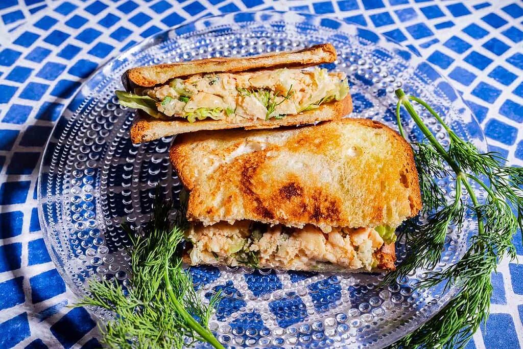 A toasted butter bean salad sandwich filled with a chunky mixture, lettuce, and herbs sits on a clear plate, garnished with sprigs of fresh dill, on a blue patterned tablecloth.