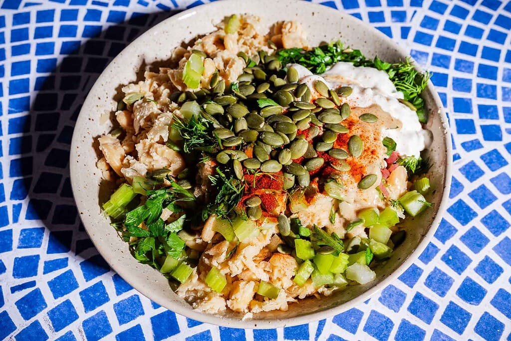 A bowl filled with chopped vegetables, herbs, seeds, and a creamy sauce—perfect as a vibrant butter bean salad sandwich filling—served on a blue and white mosaic-patterned surface.