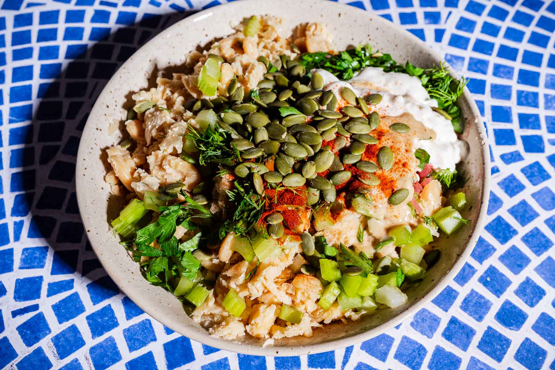 A bowl filled with chopped vegetables, herbs, seeds, and a creamy sauce—perfect as a vibrant butter bean salad sandwich filling—served on a blue and white mosaic-patterned surface.