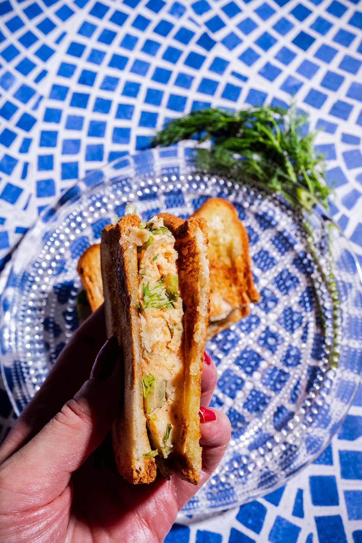 A hand holds a toasted butter bean salad sandwich with flaky, herbed fish filling, over a clear glass plate on a blue and white tiled surface.
