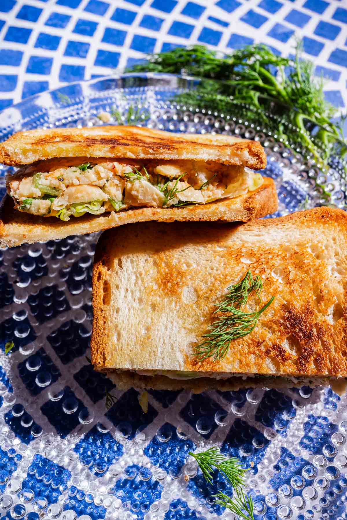 A toasted butter bean salad sandwich, cut in half and filled with chopped ingredients and fresh herbs, is placed on a clear glass plate with a sprig of dill, set on a blue and white mosaic surface.