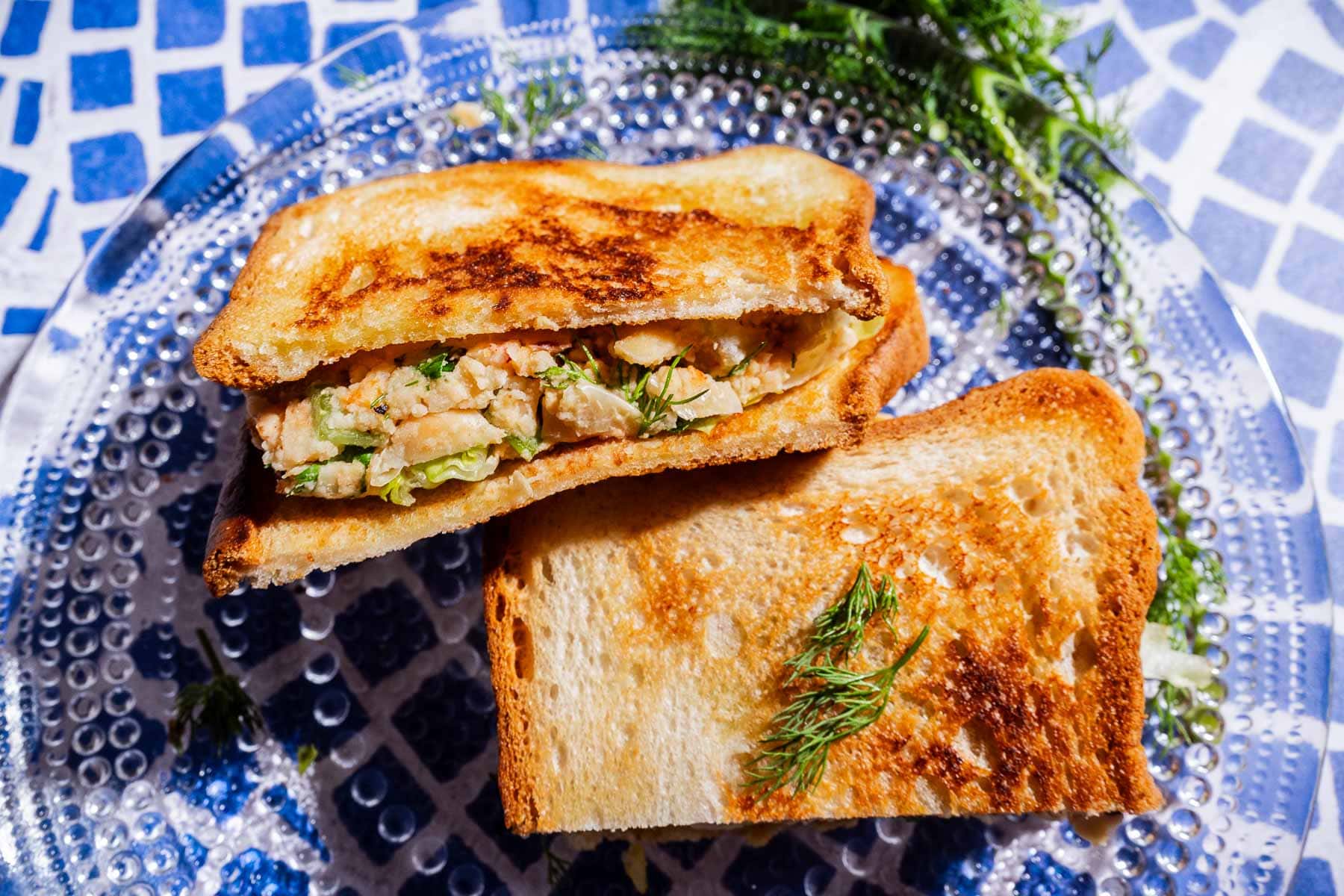 A toasted butter bean salad sandwich filled with a creamy chicken and herb mixture, garnished with fresh dill, sits on a clear glass plate over a blue and white tiled surface.