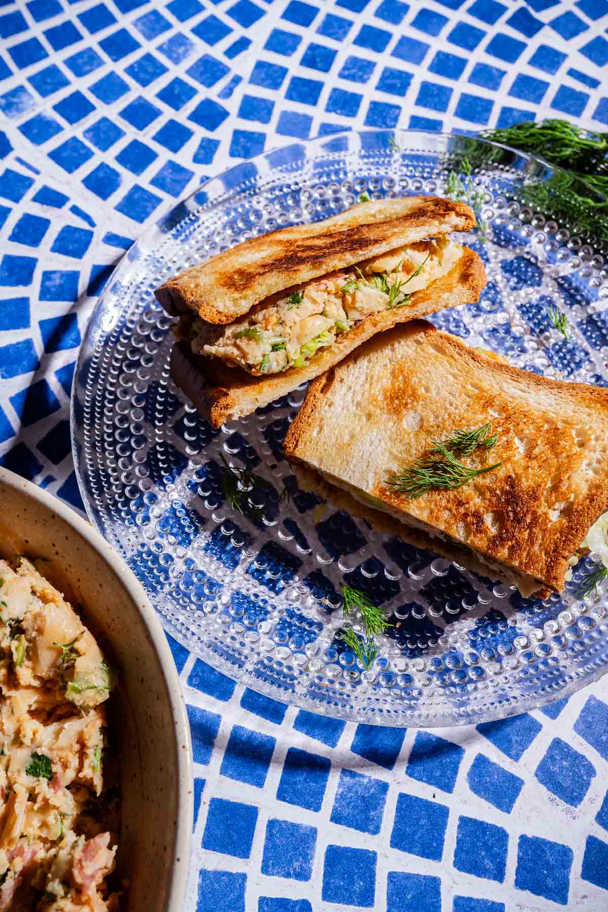 Two toasted butter bean salad sandwiches filled with a creamy mixture, garnished with dill, are served on a clear glass plate atop a blue and white tiled surface.