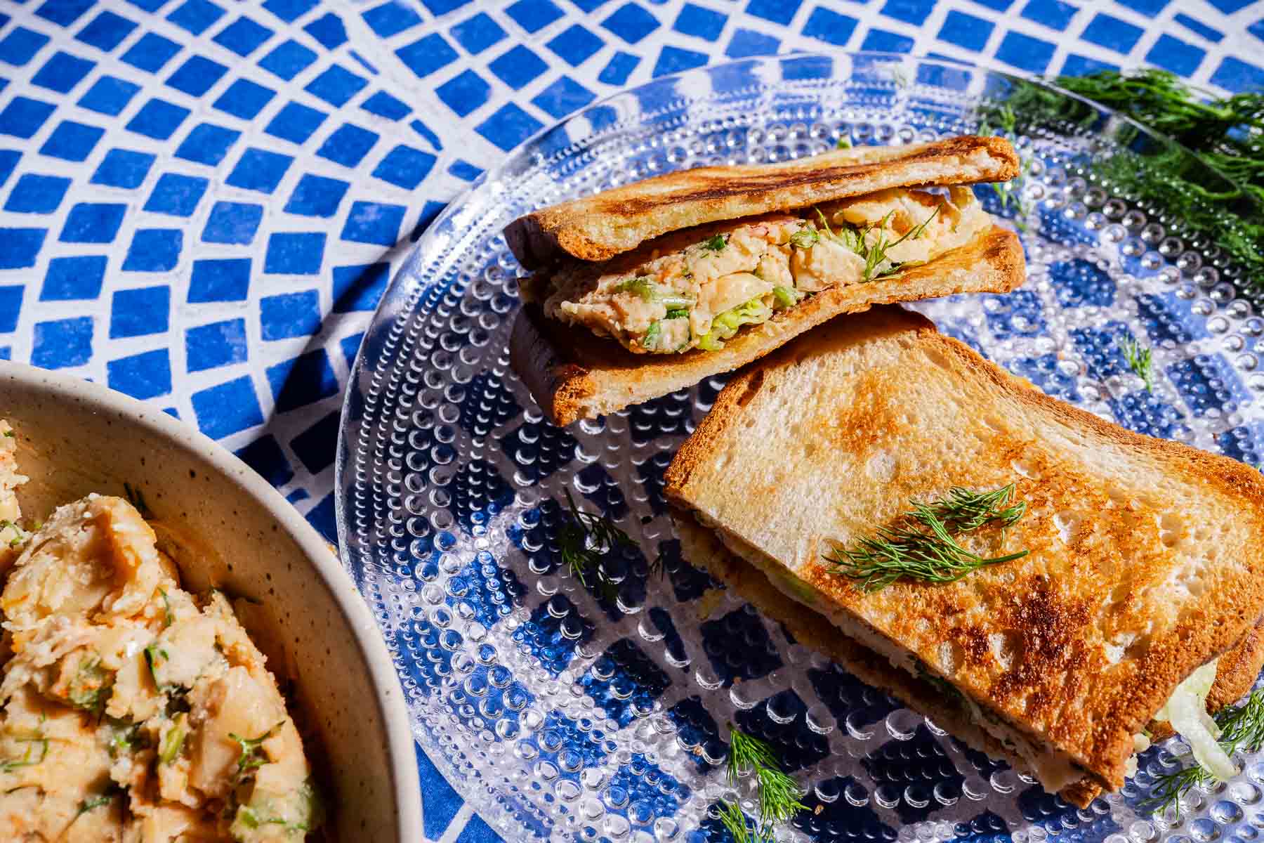 A butter bean salad sandwich, filled with a chunky, creamy mixture and fresh dill, sits on a glass plate atop a blue and white mosaic table.