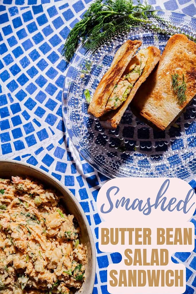 A smashed butter bean salad sandwich with fresh dill on toasted bread, served on a clear plate next to a bowl of butter bean salad, rests atop a blue patterned surface.