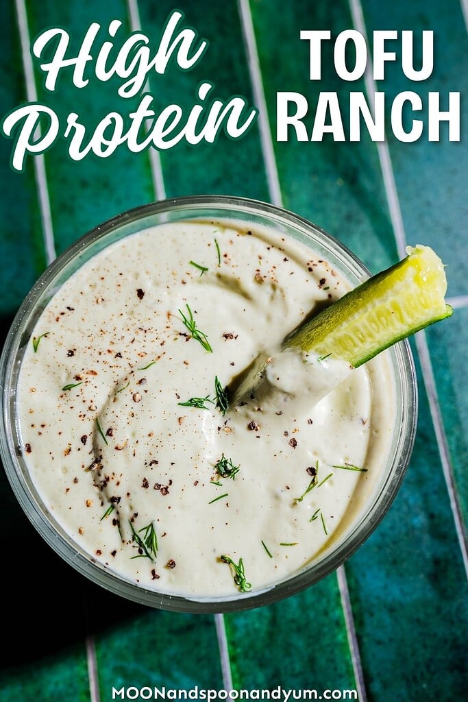 A glass bowl of creamy tofu ranch dip garnished with fresh herbs, black pepper, and a cucumber stick sits on a green striped surface. Text reads: "High Protein Tofu Ranch." Perfect for dipping and packed with plant-based protein.