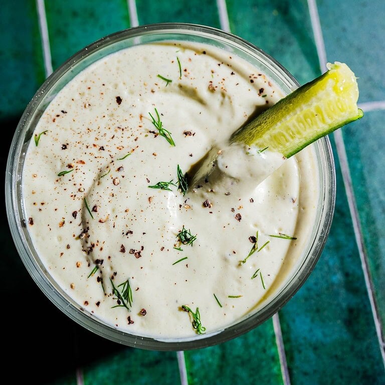 A bowl of creamy tofu ranch dip garnished with fresh dill and black pepper, with a cucumber spear partially submerged, sits on a green tiled surface.