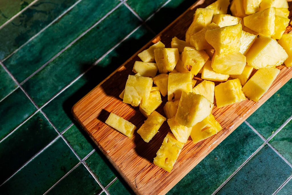 Chunks of fresh pineapple are arranged on a wooden cutting board set on a green tiled surface.
