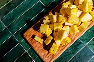 Chunks of fresh pineapple are arranged on a wooden cutting board set on a green tiled surface.