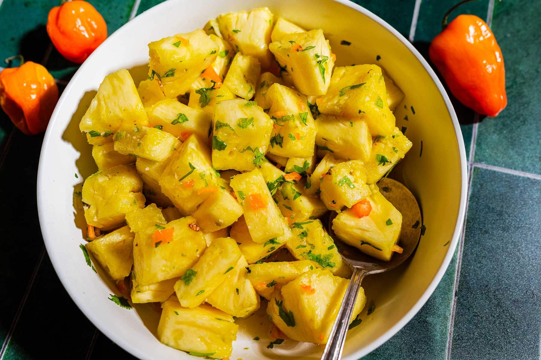 A bowl of chopped pineapple mixed with herbs and small orange pepper pieces, with a spoon inside. Two whole orange peppers are placed next to the bowl on a green tiled surface.