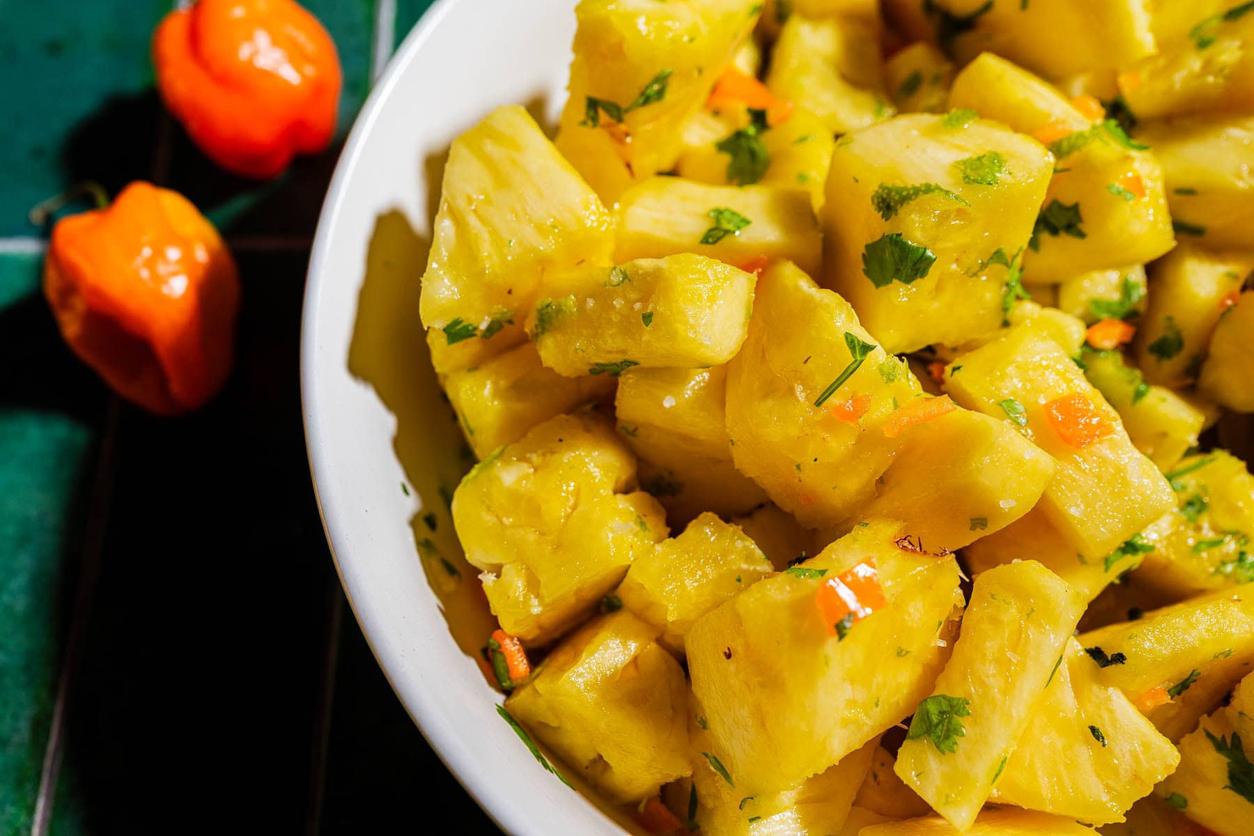 A white bowl filled with pineapple chunks mixed with herbs and diced peppers sits on a green tiled surface with two orange peppers nearby.