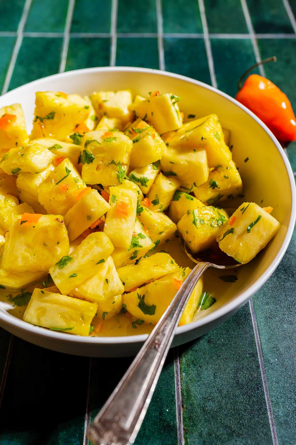 Bowl of cubed yuca salad mixed with chopped herbs and orange peppers, with a fork resting inside the bowl on a green tiled surface.