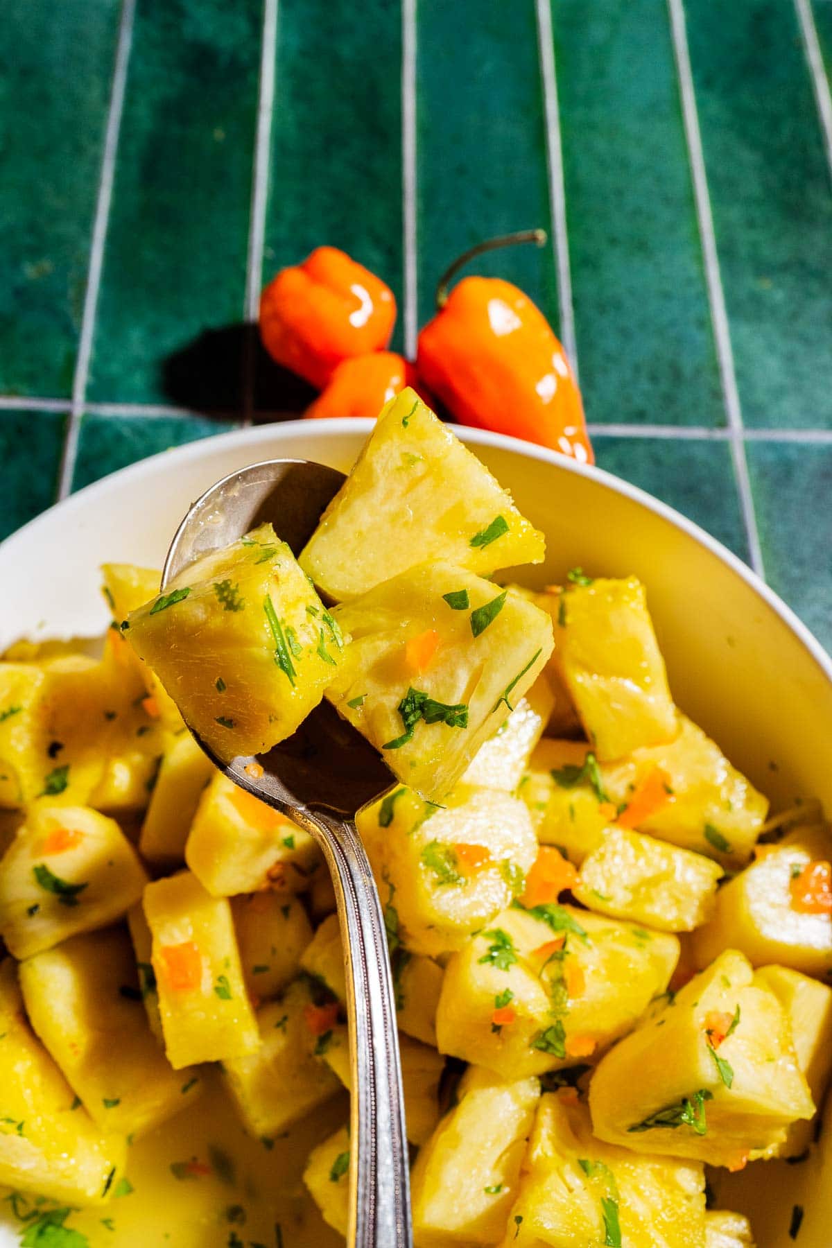 A spoonful of cooked, seasoned yellow yam pieces with herbs, held above a bowl of more yam cubes. Two orange peppers are placed on the green tiled surface in the background.