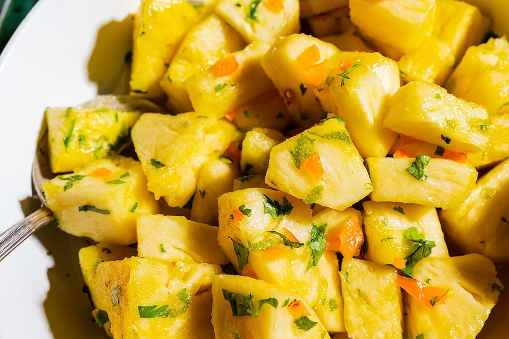 A bowl of chopped pineapple pieces mixed with herbs and small orange pepper pieces, with a metal spoon on the side.