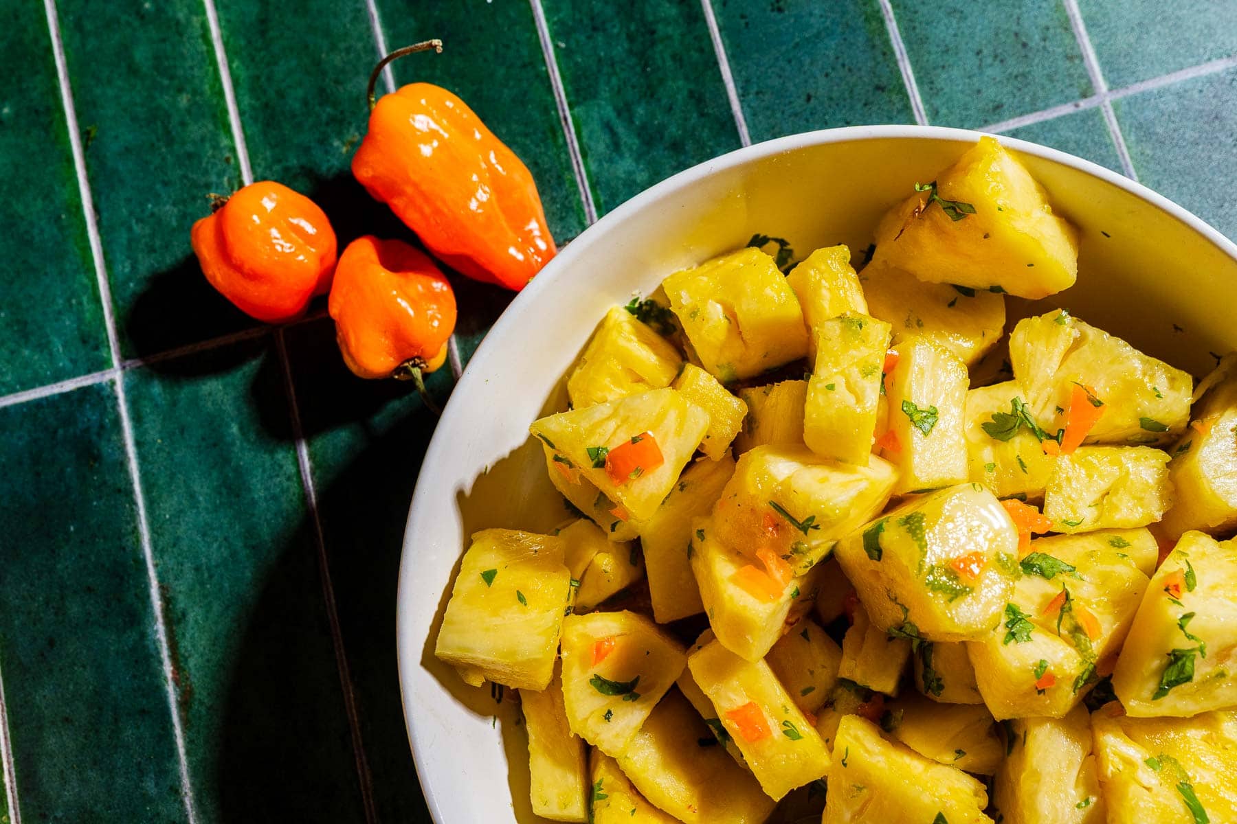Bowl of diced, seasoned yuca garnished with herbs, next to two orange habanero peppers on a green tiled surface.