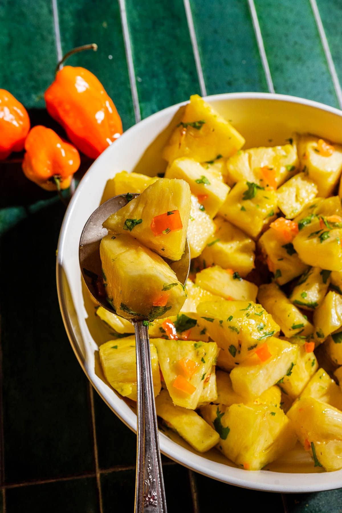 A bowl of pineapple chunks mixed with herbs and small orange pepper pieces, with a spoon lifting some pieces, sits on a green tiled surface next to two whole orange peppers.