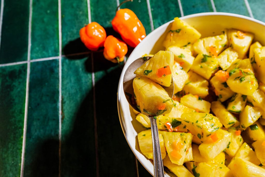 A bowl of pineapple salad with herbs and chopped peppers sits on a green tiled surface, with a spoon and three whole orange peppers nearby.