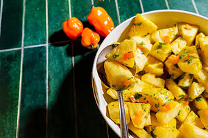 A bowl of pineapple salad with herbs and chopped peppers sits on a green tiled surface, with a spoon and three whole orange peppers nearby.