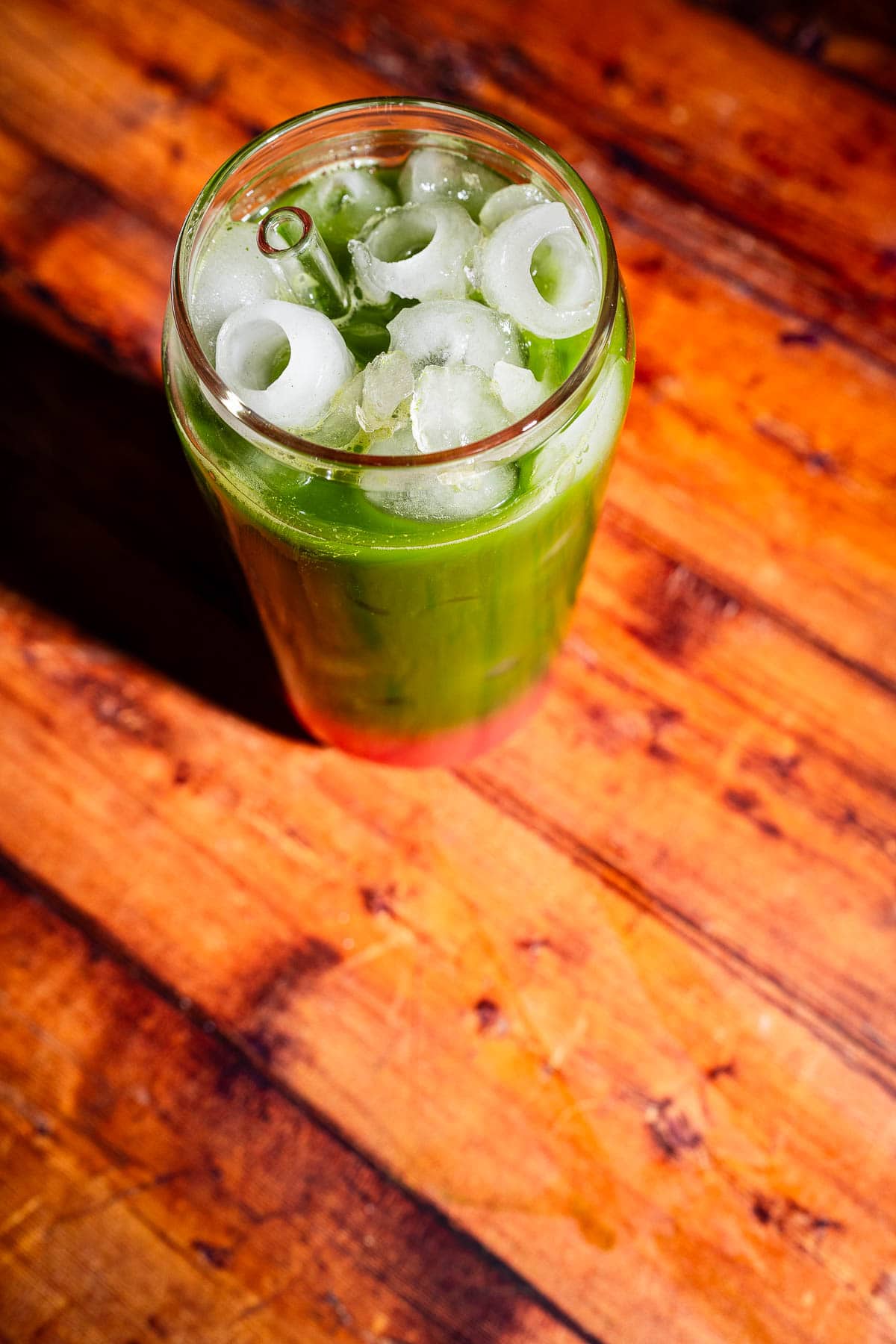 A glass of iced green beverage with round ice cubes sits on a wooden surface in direct sunlight.